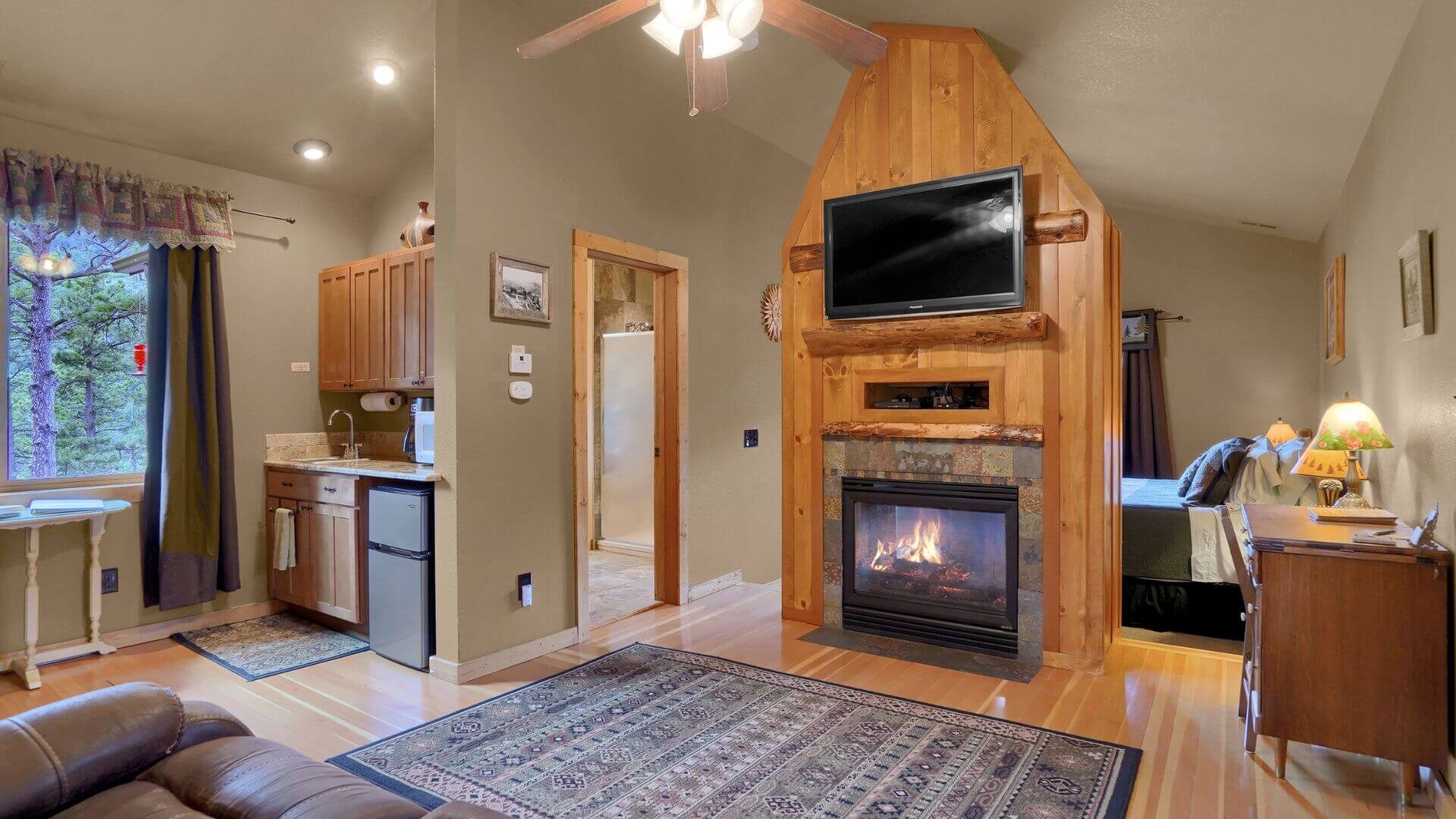 An open floor plan living room with a fireplace, flat screen TV, writing desk, a kitchen area, and a doorway to a bedroom with a king bed