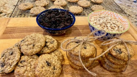 A board and cooling rack full of oatmeal raisin cookies, and bowls of oats and raisins next to them.
