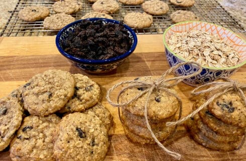 A board and cooling rack full of oatmeal raisin cookies, and bowls of oats and raisins next to them.